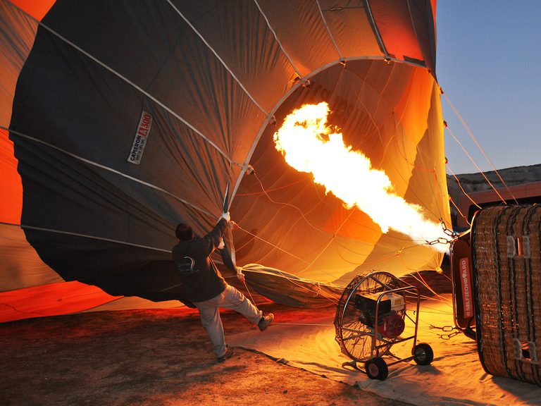 Crew inflating a balloon envelope on the ground with a long burner flame at dawn