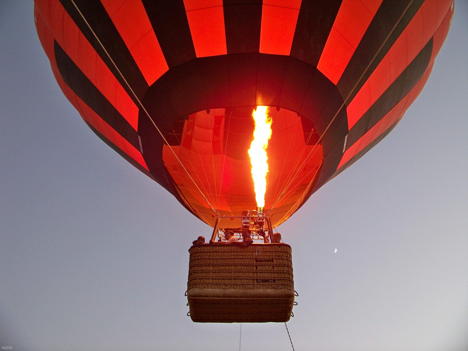 Red and black hot air balloon overhead with the burner lit against a pale sky